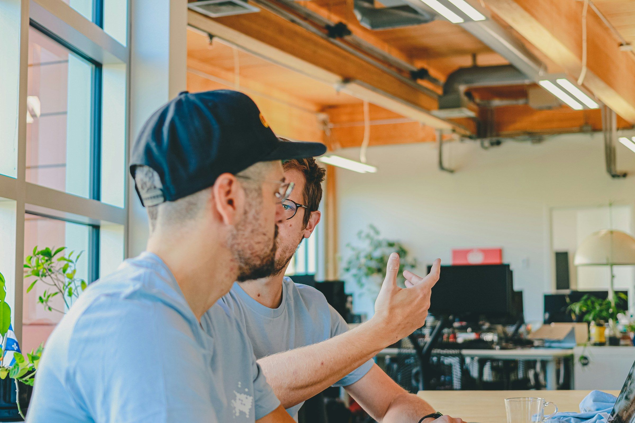 Two colleagues talk in an open-plan office; one explains with a gesture of the hand.