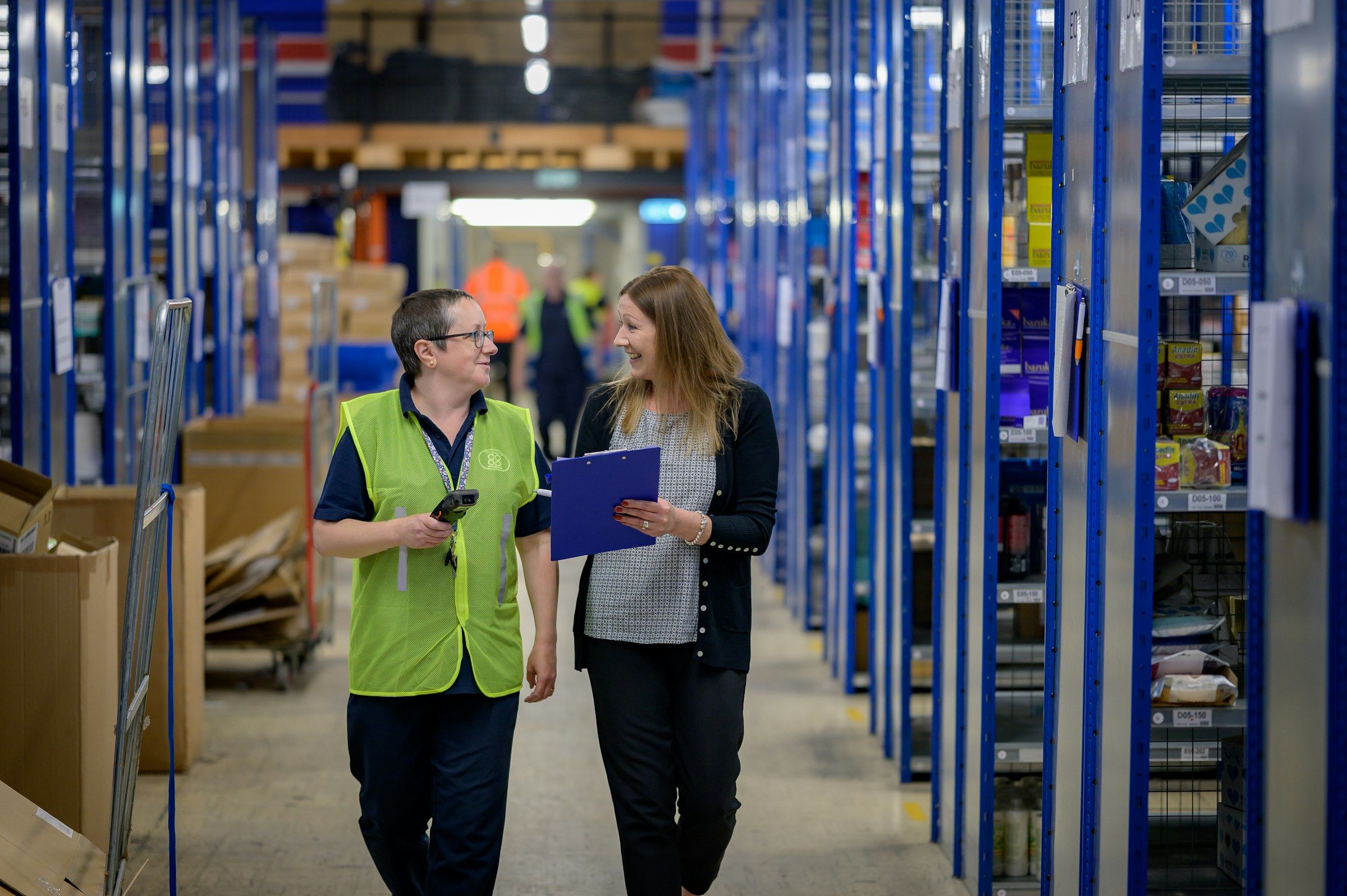 Two colleagues walk and talk in a warehouse aisle; one holds a clipboard.