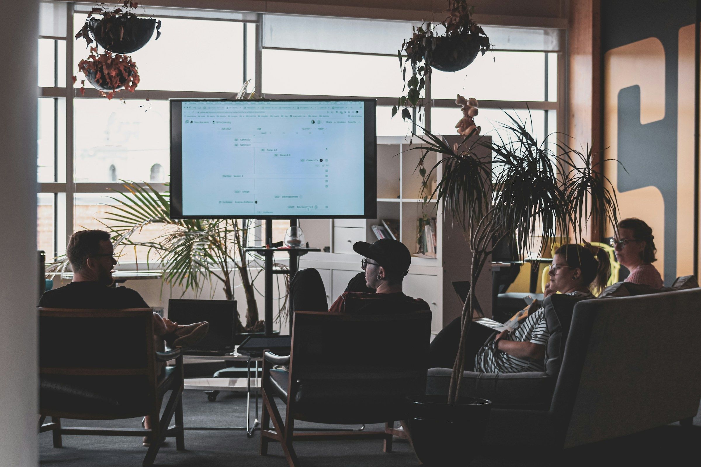 In a bright room, a team seated in a half-circle follows what appears on a large screen.